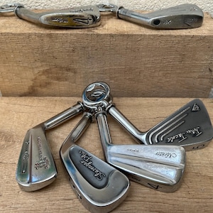 May include: A collection of six silver-toned metal golf club heads repurposed as bottle openers. Each club head features engraved brand names including Wilson, Slazenger Plus, Mentor, and John Jacobs, with visible club numbers and forged markings.