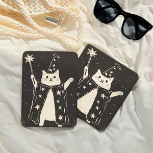 Puede incluir: Dos posavasos rectangulares negros con una ilustración blanca de un gato mago sosteniendo una varita de estrella. El gato lleva un sombrero y una capa de mago con estrellas. Gafas de sol y una bolsa de red crema en el fondo.