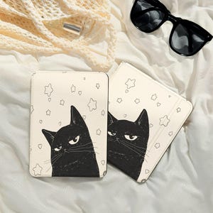 May include: Two cream-colored tablet cases with a black cat illustration and star and heart doodles. The cat has a grumpy expression. Black sunglasses and a cream-colored bag are in the background.