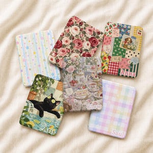 Peut inclure: Cinq étuis rectangulaires pour liseuse avec des motifs variés : rayures et étoiles pastel, roses rouges et roses, patchwork avec un chiot, chat noir dans une piscine, et un motif à carreaux pastel avec le nom Saul inscrit en bas.