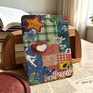 May include: A rectangular e-reader cover featuring a collage of patterns and illustrations including a yellow star, a red apple with a heart-shaped bite, a white cat wearing sunglasses, a blue butterfly, a sunflower, and the name Perciv in white text.
