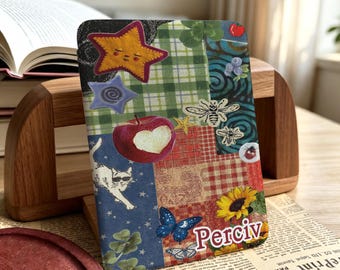 May include: A rectangular e-reader cover featuring a collage of patterns and illustrations including a yellow star, a red apple with a heart-shaped bite, a white cat wearing sunglasses, a blue butterfly, a sunflower, and the name Perciv in white text.