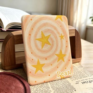 May include: A rectangular e-reader case featuring a hand-painted swirling peach and cream background with yellow stars and small orange dots. The name Pearl is printed in white script lettering in the bottom right corner of the device cover.