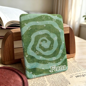 May include: A rectangular e-reader cover featuring a green and light green swirling spiral pattern with white star outlines and small white dots. The name Fiona is printed in white letters in the bottom right corner of the cover.