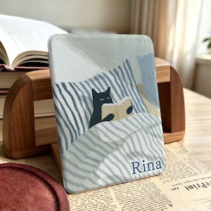 May include: A rectangular e-reader cover featuring a graphic illustration of a black cat reading a book while tucked under blue and white striped bedding. The name Rina is printed in dark blue letters at the bottom right corner of the cover.