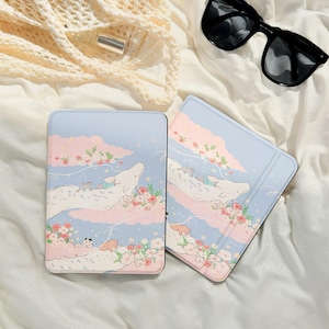 Könnte beinhalten: Zwei hellblaue Tablet-Hüllen mit einem skurrilen Design. Die Hüllen zeigen ein weißes Wesen, das durch rosa Wolken schwebt, umgeben von Blumen und kleinen Katzen. Schwarze Sonnenbrillen sind im Hintergrund.