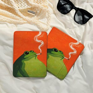 May include: Two rectangular coasters with an orange background feature a green frog smoking a cigarette. The frog has large black eyes and white smoke curls upwards. The coasters are on a white surface.
