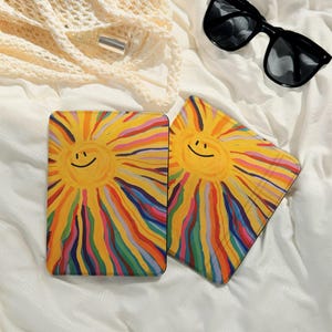 May include: Two rectangular coasters with a smiling sun design. The sun is yellow with colorful rainbow rays. The coasters are on a white surface with a pair of black sunglasses and a beige bag.