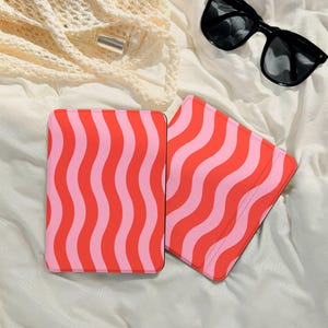 Puede incluir: Dos estuches rectangulares con un patrón ondulado de rayas rojas y rosas alternas. Los estuches están sobre una superficie blanca, con gafas de sol negras y una bolsa de red beige en el fondo.