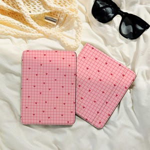 Puede incluir: Dos cuadernos rosas con un patrón de cuadrícula y pequeños corazones rojos. Los cuadernos son rectangulares con un borde negro. Gafas de sol negras y una bolsa color crema en el fondo, sobre una superficie blanca.