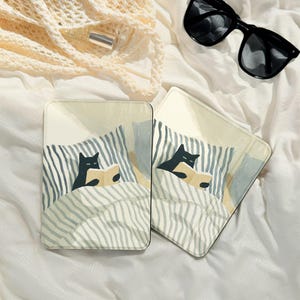 Puede incluir: Dos posavasos cuadrados con una ilustración caprichosa de un gato negro leyendo en la cama. El diseño presenta una manta a rayas en tonos grises y blancos, sobre un fondo beige claro. Gafas de sol y una pequeña bolsa en el fondo.