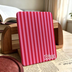 May include: A rectangular e-reader cover featuring a vertical striped pattern in alternating shades of bright red and light pink. The name Rina is printed in white, stylized serif lettering with a thin red outline in the bottom right corner.
