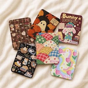 Può includere: Cinque custodie rettangolari con diversi design grafici: un motivo a quadri marroni con orsi, una scena con zucche e fantasmi e la scritta 'Happy Halloween!', un coniglio con 'Bunny Bunny' e 'Anna', una stampa nera con gatti, un motivo trapuntato e un arcobaleno floreale con 'Saul'.