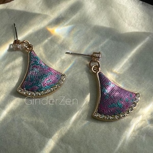 Puede incluir: Un par de pendientes dorados con un diseño en forma de abanico. Los pendientes presentan una incrustación de tela morada y verde azulado, delineada con pequeños cristales transparentes. Cierre de poste.