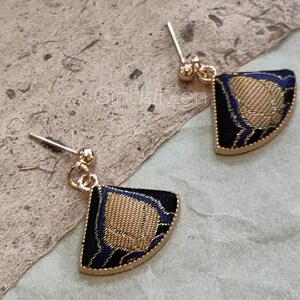 Puede incluir: Un par de pendientes de botón dorados. Cada pendiente presenta un diseño en forma de abanico con fondo negro, centro dorado y detalles azules. Los pendientes tienen un borde dorado y un poste para orejas perforadas.