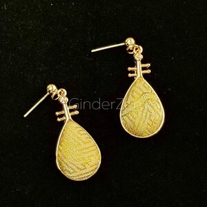 May include: A pair of gold-coloured earrings with a teardrop-shaped design. The earrings feature a textured, yellow-gold interior and a decorative top, resembling a musical instrument. The earrings are displayed against a black background.