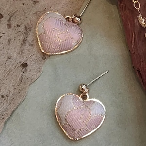 Puede incluir: Un par de pendientes en forma de corazón. Cada pendiente presenta un corazón estampado en rosa y blanco con un borde dorado. Los pendientes tienen un cierre de poste y bola para orejas perforadas. El fondo es de color neutro.