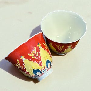 May include: Two decorative ceramic cups with a red exterior and a white interior. The cups feature a yellow and blue floral design. The rims of the cups have a scalloped edge.