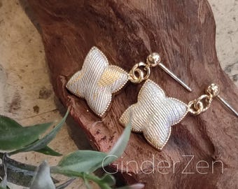 May include: A pair of gold and silver-colored earrings. Each earring features a four-petal flower design with a textured silver finish and gold accents. The earrings are on a wooden surface with the text "GinderZen".