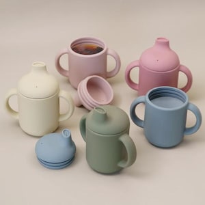 May include: A selection of pastel-coloured baby sippy cups. The cups are in shades of pink, blue, green, and cream. Some have lids and handles, while others are open. The cups are made of a soft, matte material.