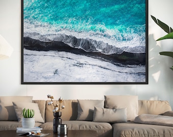 May include: A framed aerial photograph of a turquoise ocean with white waves crashing against a black and white shoreline. The artwork is displayed above a beige sectional sofa in a living room setting.