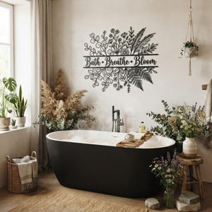 May include: A black metal wall art piece featuring a botanical design of leaves, flowers, and ferns with the text "Bath • Breathe • Bloom" centered horizontally. The intricate cutout silhouette is mounted on a plain wall above a bathtub.
