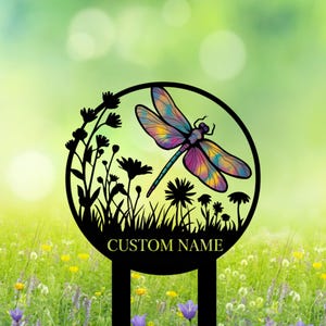 Puede incluir: Una estaca de jardín de metal negro con un diseño de libélula colorida. La libélula tiene alas multicolores y un cuerpo azul. La estaca incluye el texto "CUSTOM NAME" y está rodeada de flores y hierba estilizadas.