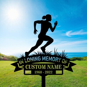 Puede incluir: Estaca conmemorativa de metal negro con la silueta de una figura corriendo. La estaca incluye el texto "IN LOVING MEMORY OF", espacio para un nombre personalizado y los años 1960-2022. Sobre un fondo de hierba verde, cielo azul y océano.