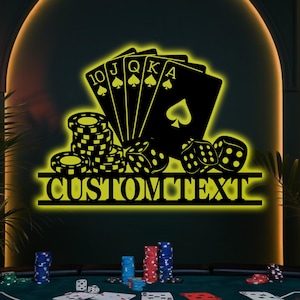 Op de afbeelding: Zwarte metalen wandkunst met een pokerthema, inclusief speelkaarten, dobbelstenen en fiches. De kaarten tonen een royal flush in schoppen. De tekst "CUSTOM TEXT" staat eronder. De kunst is verlicht met een gele gloed.