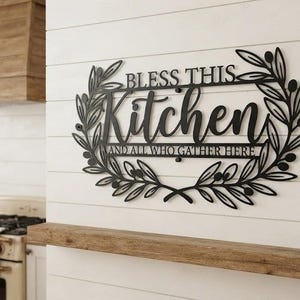 Może przedstawiać: Czarny metalowy napis ścienny z tekstem "BLESS THIS Kitchen AND ALL WHO GATHER HERE" wykonanym mieszanką czcionek szeryfowych i pisanych. Litery są otoczone symetrycznym wzorem gałązek oliwnych z liśćmi oraz drobnymi owocami oliwek.