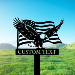 Puede incluir: Letrero de jardín de metal negro con un águila calva con las alas extendidas frente a una bandera estadounidense. El letrero incluye el texto "CUSTOM TEXT" en una base rectangular, sobre un fondo de hierba verde y cielo azul.