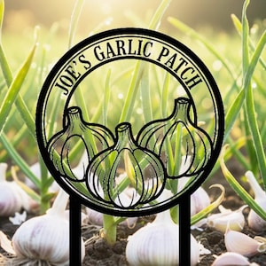 May include: A black metal garden stake with the text "JOE'S GARLIC PATCH" in a circular design, featuring three garlic bulb illustrations. The stake is placed in a garden bed with garlic plants and bulbs.