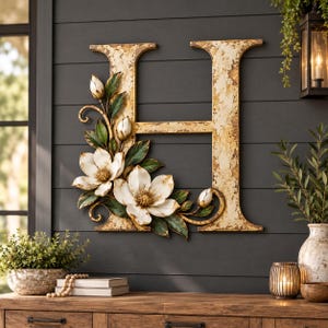 May include: A large, distressed gold-toned letter H wall decoration adorned with sculpted white magnolia flowers and green leaves. The letter features a weathered, textured finish, and the floral arrangement is attached to the left side.