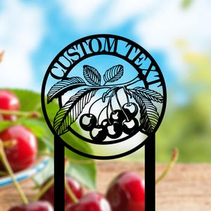 May include: Black metal cherry-themed garden stake with the words "CUSTOM TEXT" in a circle. The stake features a detailed cutout design of cherries and leaves. The background is blurred, with red cherries and a blue sky.