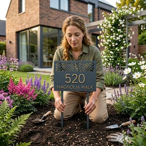 May include: A rectangular dark gray metal garden sign featuring the number 520 and the text MAGNOLIA WALK in raised gold lettering. The sign has a decorative geometric lattice pattern at the top and is supported by two thin metal stakes.