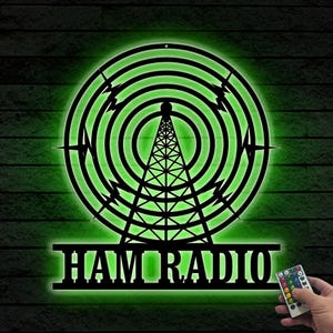 以下が含まれることがあります： 緑色のネオンの光を放つ黒い金属製のHAM RADIOサイン。サインの中央には電波塔があり、同心円と稲妻のデザインで囲まれています。「HAM RADIO」の文字が下に表示されています。