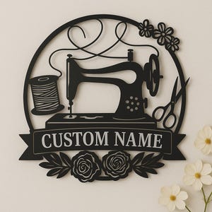 May include: Black metal sewing machine wall art featuring a spool of thread, scissors, and floral details. The design includes a banner with the text "CUSTOM NAME" and is set within a circular frame.