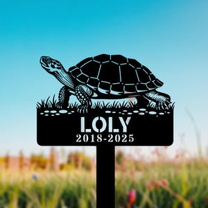 May include: Black metal garden stake featuring a turtle design. The stake includes the name "LOLY" and the years "2018-2025". The turtle is depicted with detailed lines, standing on a patch of grass.
