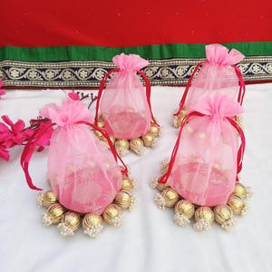 Könnte beinhalten: Vier rosa Organza-Kordelzugbeutel mit roten Bändern, jeweils auf einer goldfarbenen Perlenbasis mit weißen Perlenakzenten. Die Beutel haben ein Blumenmuster und sind vor rotem und weißem Hintergrund platziert.