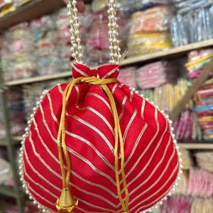 Pode incluir: Uma bolsa vermelha com cordão e listras prateadas e acabamento de pérolas. A bolsa tem um cordão dourado e borlas douradas. As alças são feitas de pérolas brancas.