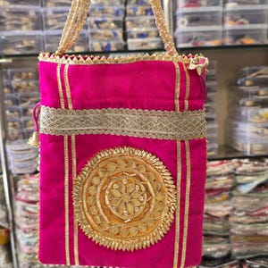 May include: A vibrant pink handbag with gold embellishments. The bag features a central circular design and gold trim. The handles are made of beaded strands. The bag is rectangular and appears to be made of fabric.