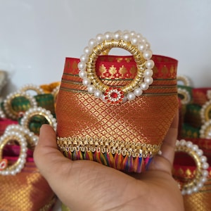 May include: A small, decorative fabric basket with a gold handle adorned with faux pearls. The basket is primarily red and gold, with intricate patterns and a red jewel. The bottom edge has a colorful fringe.