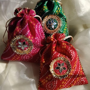 May include: Three colourful drawstring bags in red, pink, and green. Each bag features a decorative, circular embellishment with mirrored accents and gold trim. The bags have a dotted pattern and are tied with a drawstring.