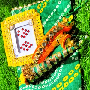 May include: An assortment of colorful Indian accessories, including a yellow-framed item with red dots, orange and gold bangles, and a green patterned pouch. The items are arranged on a bed of green grass.