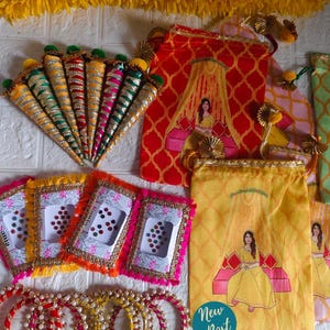 May include: A collection of colorful Indian wedding accessories. Includes decorative cones, patterned pouches with a woman illustration, framed cards with gems, and beaded bangles. The color palette features red, yellow, pink, and green.