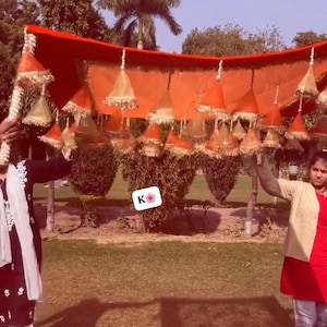 Puede incluir: Un dosel decorativo de tela naranja con borlas doradas colgantes. El dosel está sostenido por dos personas en un parque. El dosel tiene un diseño floral y probablemente se utiliza para una boda o un evento especial.