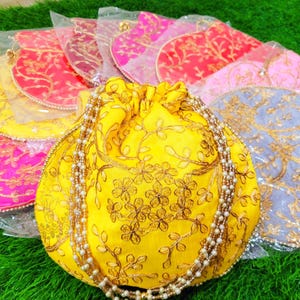 May include: A collection of colorful, embroidered drawstring bags. The central yellow bag features gold floral embroidery and a pearl beaded strap. Other bags in pink, red, and blue are visible, each with intricate designs. The bags are on a green surface.