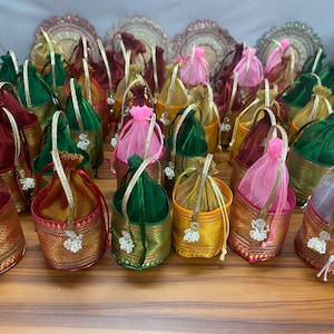 May include: A collection of colorful, ornate drawstring bags. The bags feature gold and patterned fabric bases, with various colored sheer tops in green, pink, and gold. Each bag has a decorative handle and a beaded tassel.