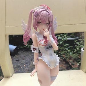 May include: A figurine of a character with pink hair in a nurse's outfit, complete with a cap and wings. The figure is holding a red object to their mouth. The outfit is white with a ruffled edge, and there is Japanese text on the chest.