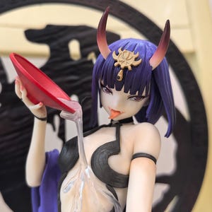 Peut inclure: Figurine détaillée d'un personnage féminin aux cheveux violets, avec des cornes rouges et un ornement de tête doré. Elle verse un liquide clair d'un bol rouge. La figurine porte un haut noir et tire la langue.